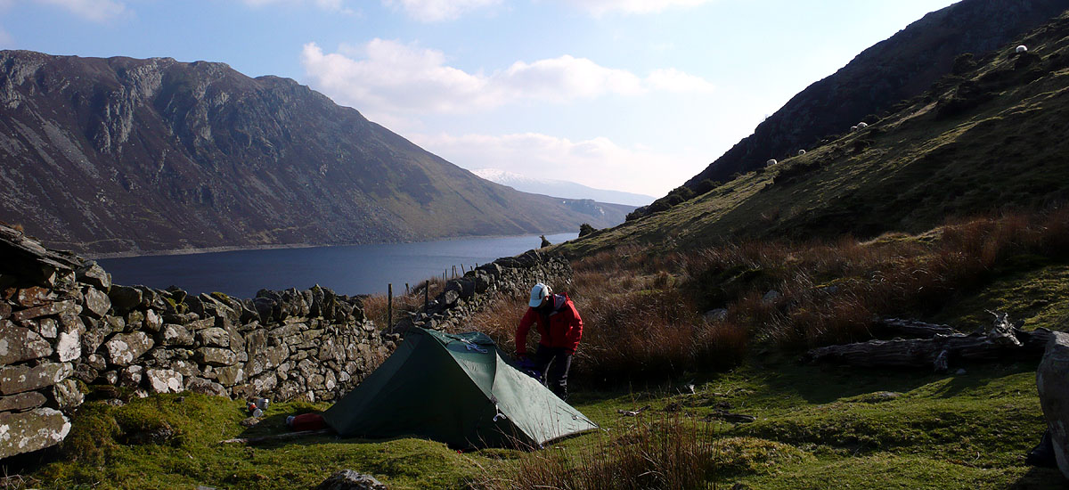 a tent sits close to a dry stoen wall, a bay in the background